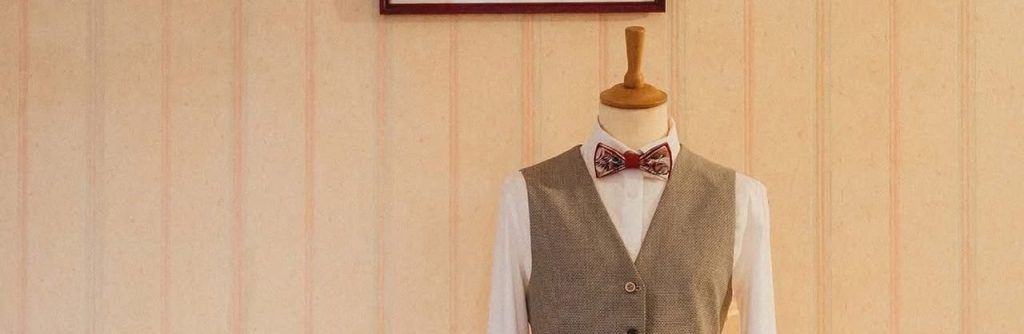 Mannequin habillé d'une chemise blanche, gilet gris et nœud papillon, sur fond rayé.
