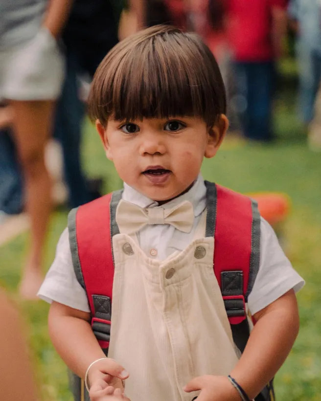 Enfant avec bretelles et nœud papillon, portant un sac à dos rouge.